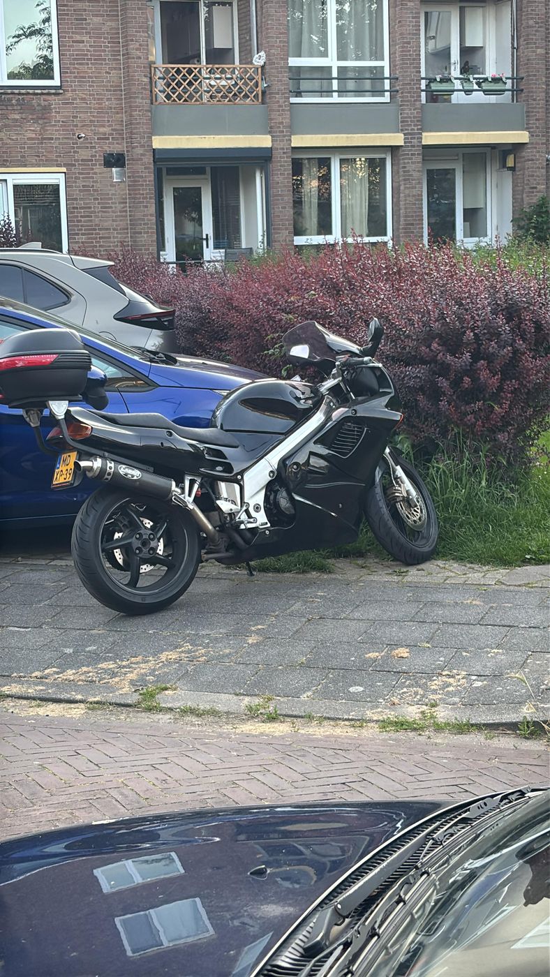 Zwarte Honda VFR-motor geparkeerd naast een blauwe auto voor een flat